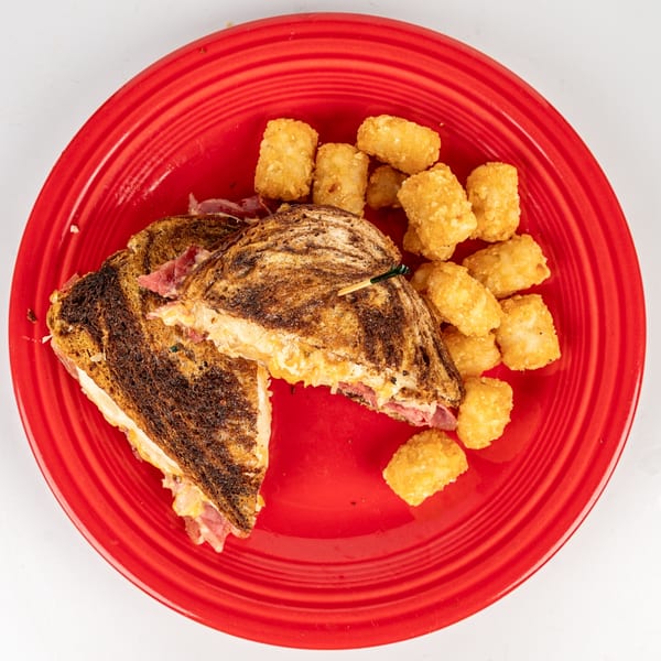 A grilled sandwich with melted cheese and meat, served with tater tots on a red plate.