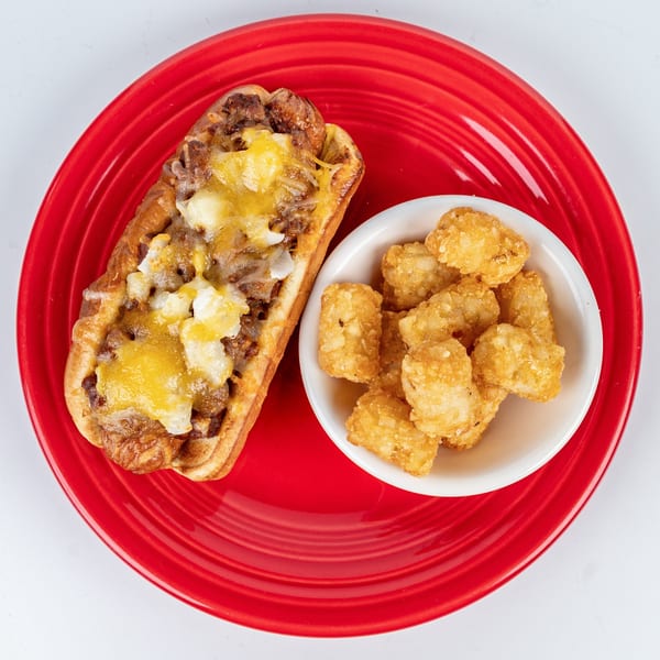 Chili cheese dog with melted cheese and a side of golden tater tots on a red plate.